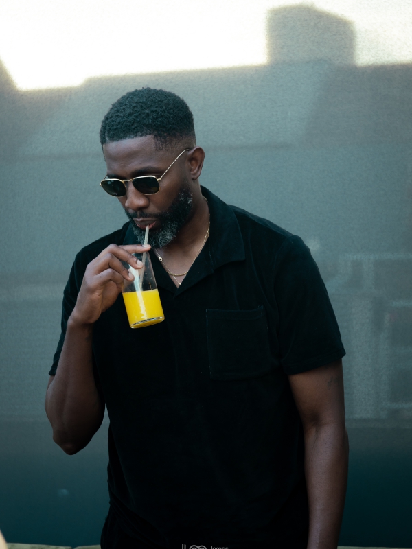 A Man In Sunglasses And A Black Shirt Drinking A Yellow Juice Through A Straw Outdoors.